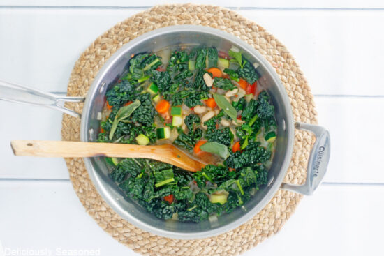 A large pan with diced carrots, kale and remaining soup ingredients in it and a wooden spoon in the pan.