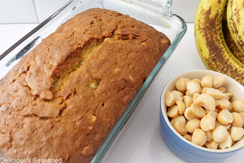 An overheat shot of a loaf of banana nut bread, bananas, and a bowl of macadamia nuts in the background. 