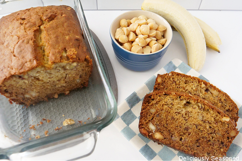 An overhead shot of banana bread on a blue and white plate, with a loaf pan, and a bowl of nuts and bananas in the background. 