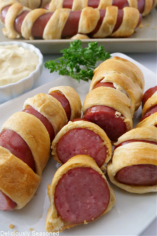 Sliced sausage wrapped in crescent rolls, laying out on a white plate with a small white bowl of dipping sauce in the background. 