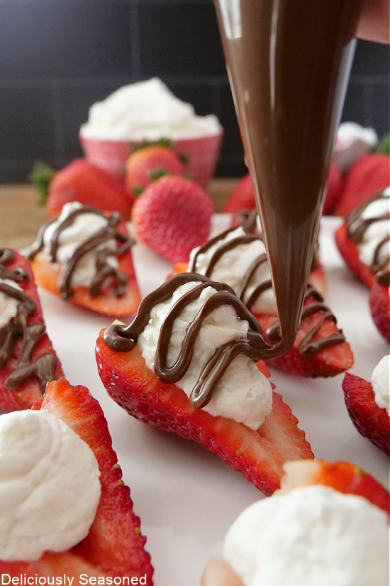 Strawberry halves, topped with whipped cream, and a piping bag full of chocolate drizzling it on top of the strawberries.