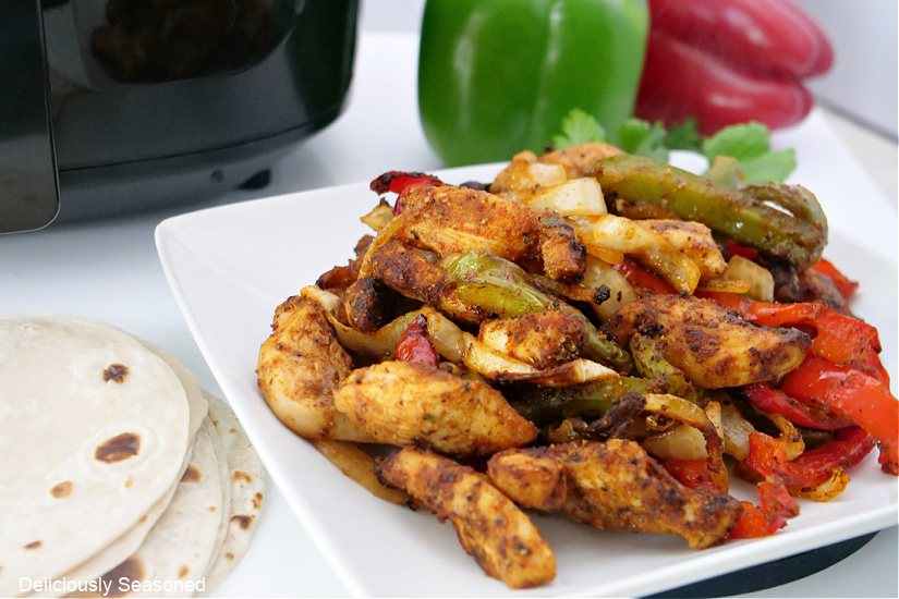 A horizontal picture of air fryer chicken fajitas on a white plate with soft, flour tortillas next to the plate and bell peppers in the background.