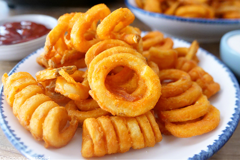 A landscape photo of a white plate with blue trim, small bowls of dipping sauces  in the background, and curly fries on the plate. 
