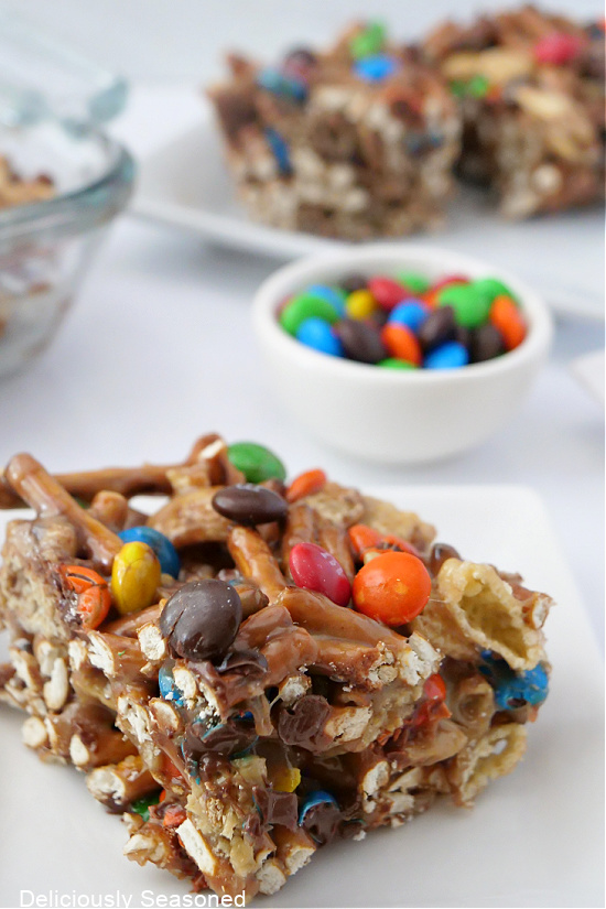 A close-up picture of a cut piece of a Chex Mix Bar on a white plate with a small bowl full of M&Ms in the background.