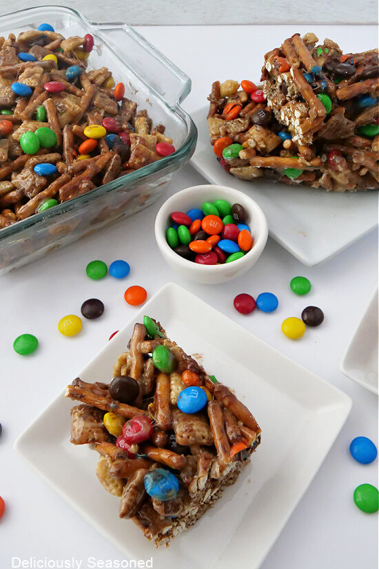 A picture of Chex Mix Bars in a glass baking dish with pieces of cut bars stacked on white plates and a small bowl of M&Ms in the middle with a few scattered around the plates.
