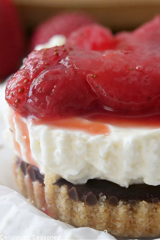 A close up photo of the edge of a mini cheesecake, showing the crust, chocolate, cheesecake, and strawberry sauce on top.