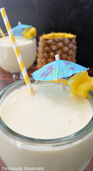 A close up photo of a pineapple milkshake in a large glass with a decorative umbrella and a yellow straw in it. 