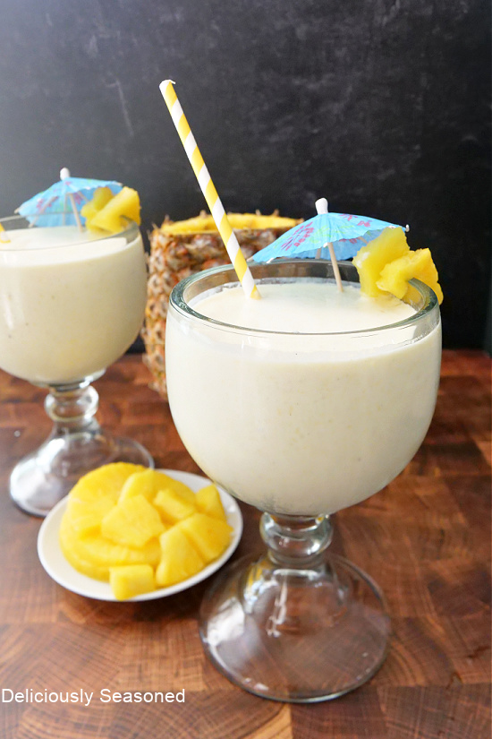 Two glasses filled with milkshakes, with a pineapple in the background and diced pineapple on a white plate.