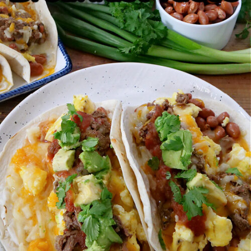 Two breakfast tacos on a white plate loaded with cilantro, salsa, beans, avocados, and green onions in the background.