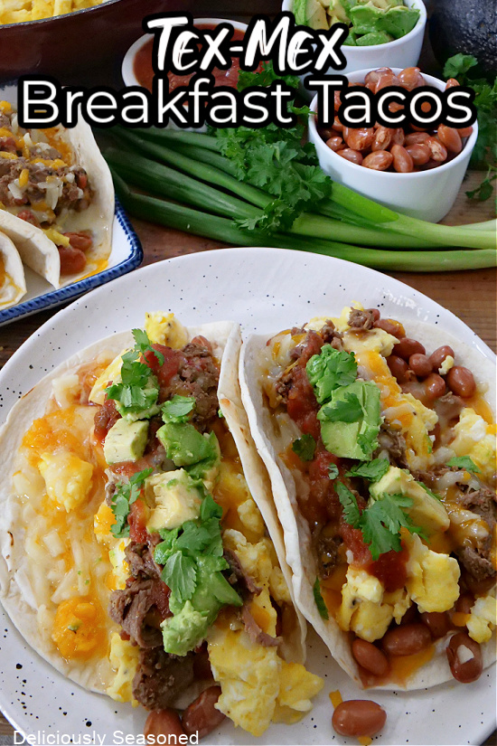 Two breakfast tacos on a white plate loaded with cilantro, salsa, beans, avocados, and green onions in the background. 