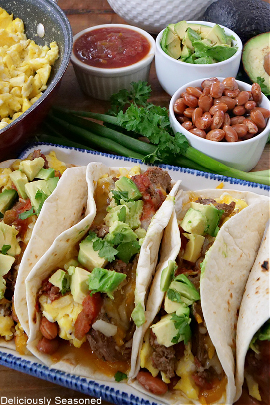 Four tacos lined up on a long white plate with a pan of scrambled eggs in the background. 
