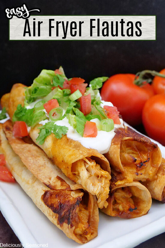 Air fryer flautas stacked on a white plate topped with sour cream, lettuce, tomatoes, and green onions.