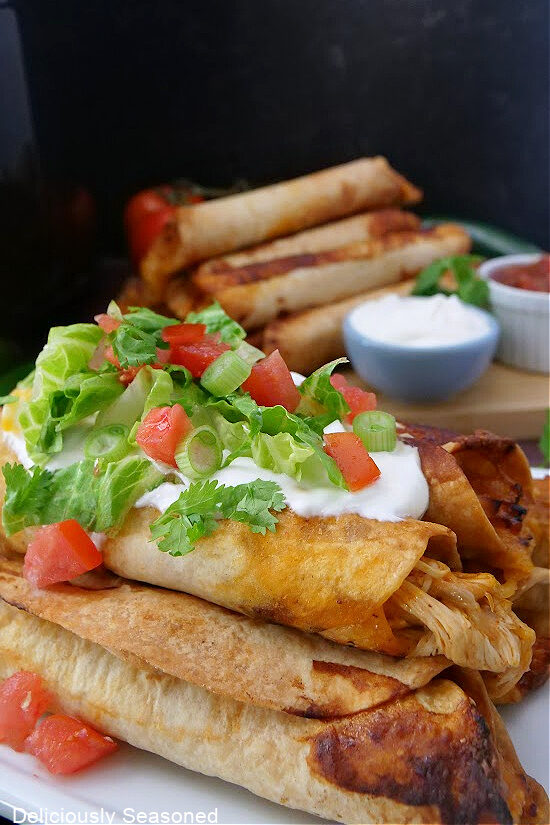 A close-up picture of Air Fryer Flautas stacked on a plate and topped with sour cream, lettuce, tomatoes, cilantro, and green onions.