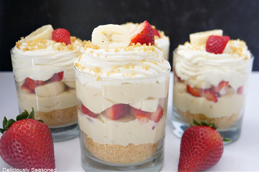 A landscape photo of banana parfaits on a white board, all sprinkled with graham cracker crumbs and slices of bananas and strawberries.