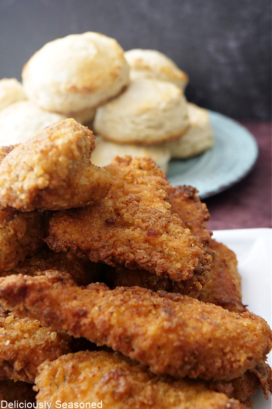 Pieces of fried chicken stacked up on a plate with biscuits stacked up in the background.