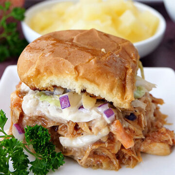 A BBQ chicken slider on a white plate with a bowl of pineapple in the background.