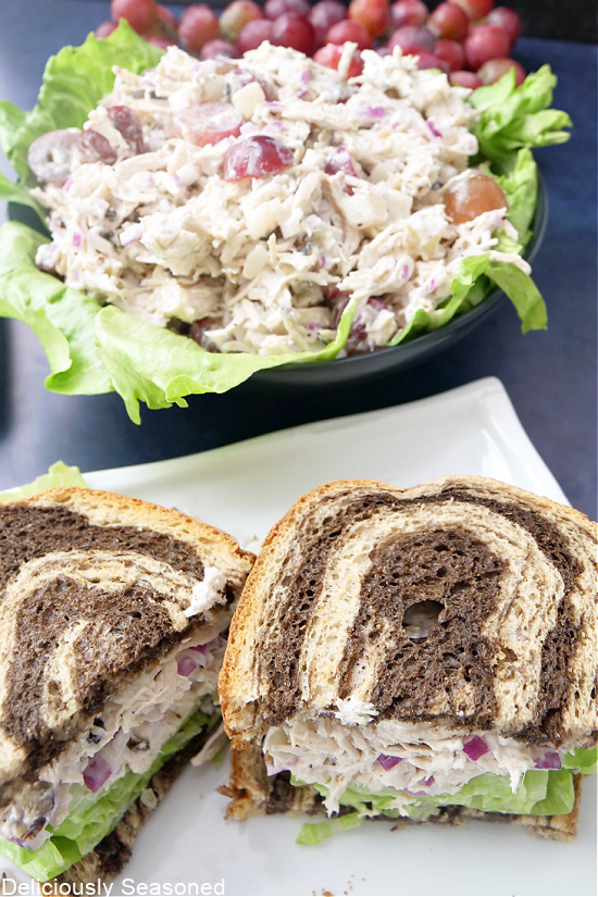 A pumpernickel chicken salad sandwich on a long white plate with a black bowl filled with chicken salad in the background.