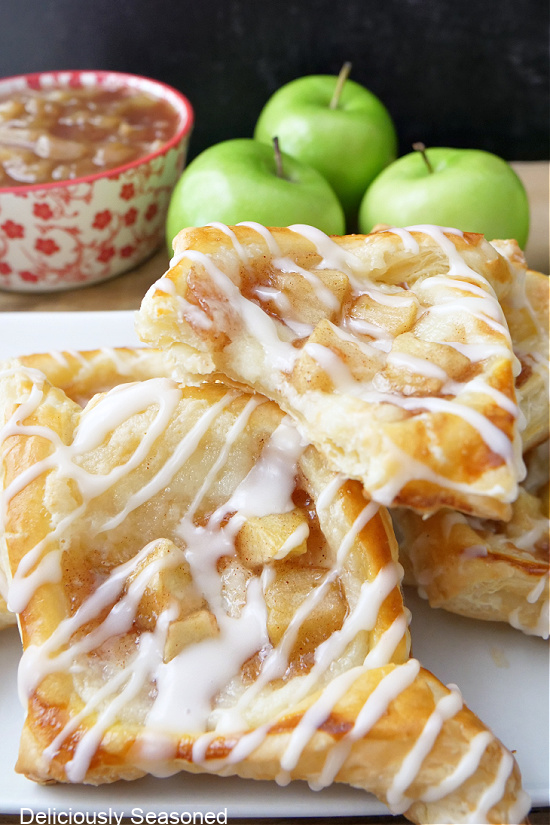A small bowl of apple pie filling in the background, next to Granny Smith apples, with puff pastries stacked on a white plate.