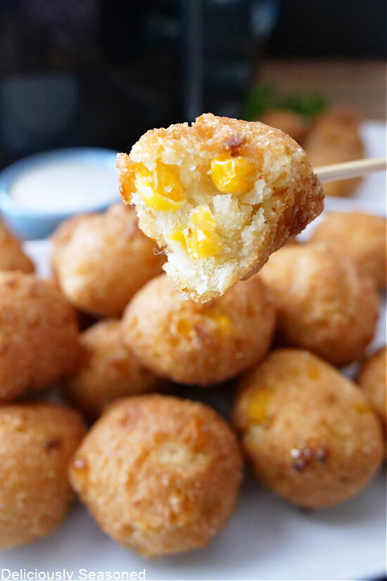 A bunch of corn hushpuppies on a white plate with one on a little skewer with a bite taken out of it.