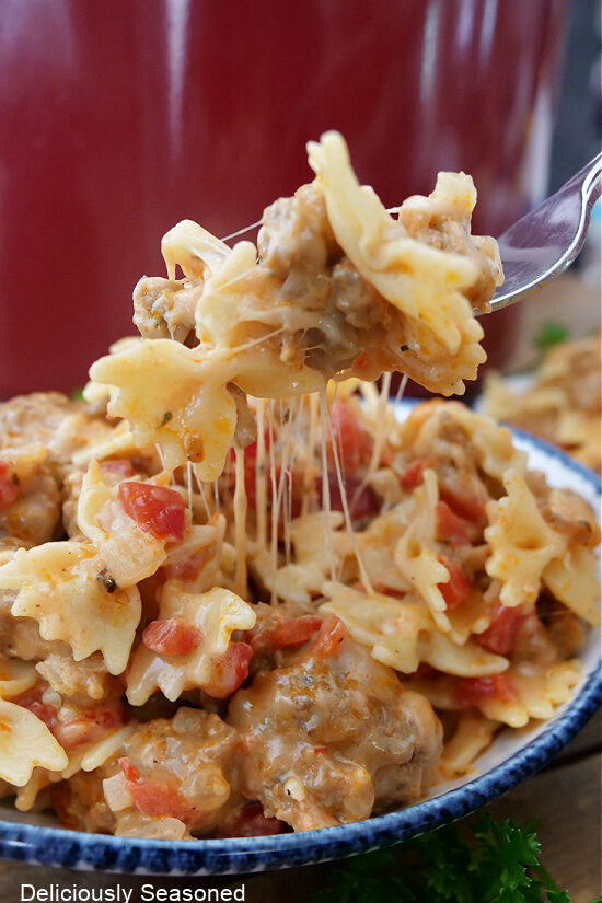 A forkful of cheesy Italian sausage bow tie pasta on a fork with a serving in a white bowl underneath.