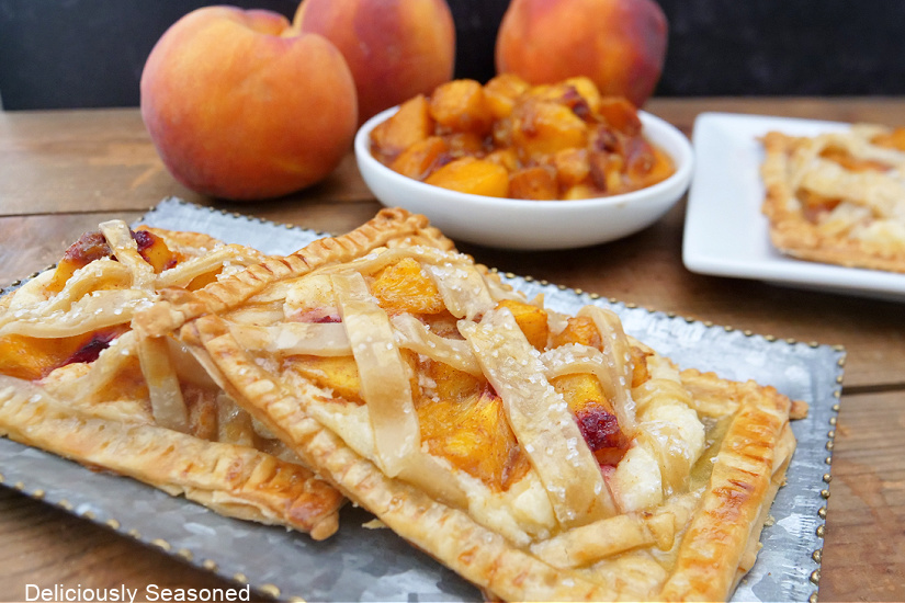 A silver tray with two peach pastries on it with fresh peaches in the background.