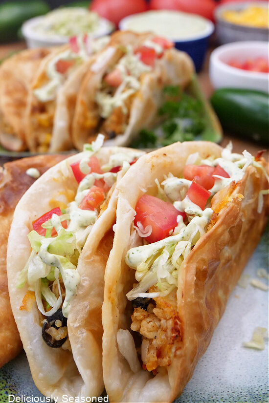Three fried chicken tacos on a plate with more tacos in the background.