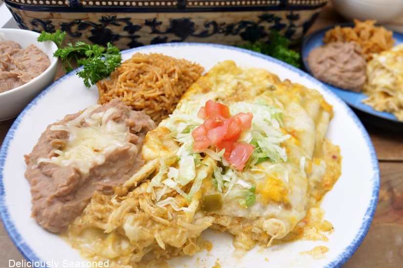 A white dinner plate with blue trim with chicken enchiladas, rice and refried beans on it.