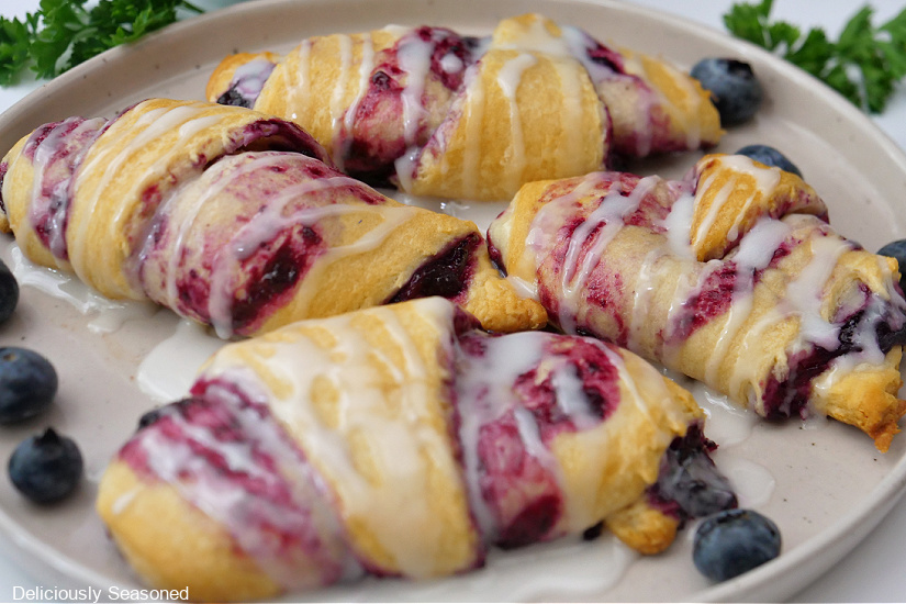 A big round plate with four crescent rolls covered in a glaze and fresh blueberries laying around the rolls.