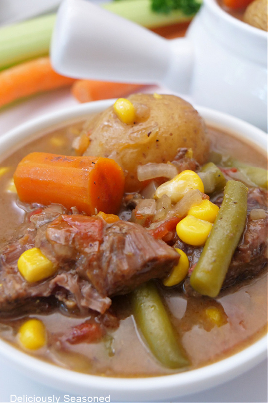 A close up photo of a bowl of beef stew that is loaded with beef, green beans, carrots, potatoes, and corn.