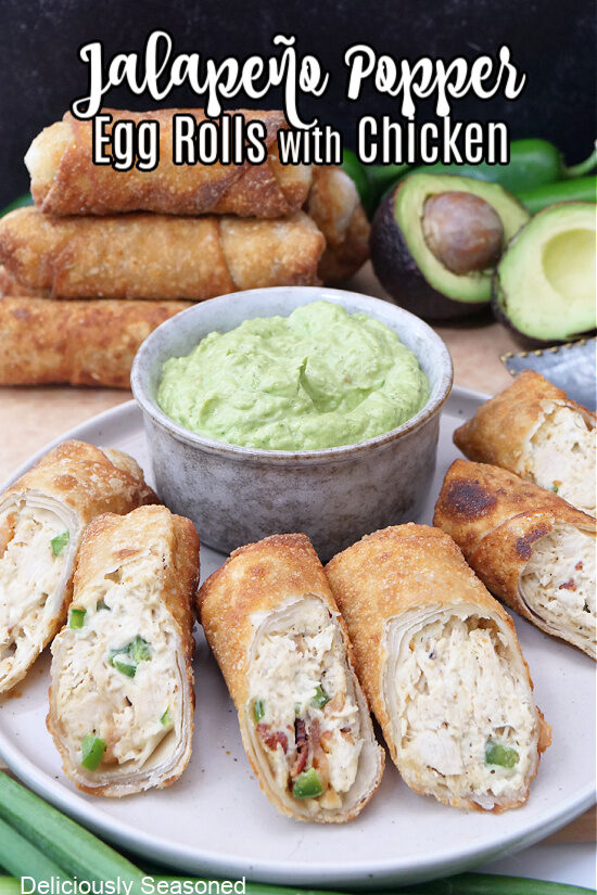 A grey round plate with 6 egg rolls on it with a grey bowl filled with avocado dip