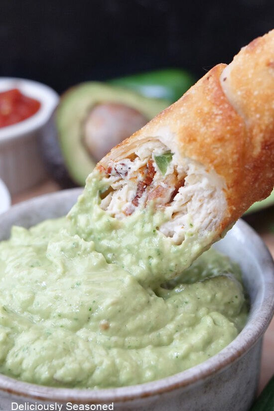 A close up of a jalapeno popper egg roll with chicken being dipped in avocado dip.