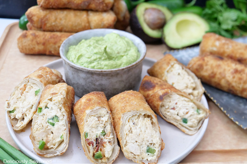 A horizontal photo of a grey plate with 6 egg rolls on it with a grey bowl filled with avocado dip and more egg rolls in the background.