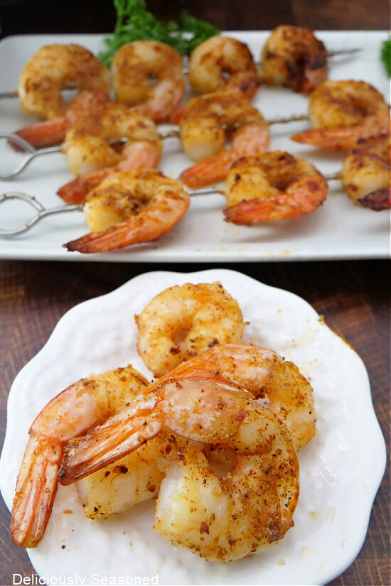 A small white plate with jumbo shrimp on it and another white plate in the background with more air fryer Cajun shrimp on skewers place on it.