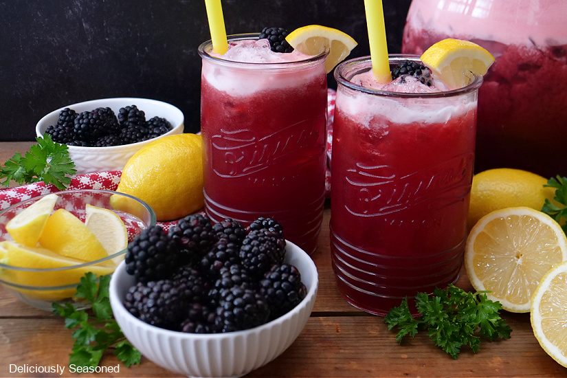 Two glasses of lemonade with two small white bowls full of blackberries and lemons laying around the cups.