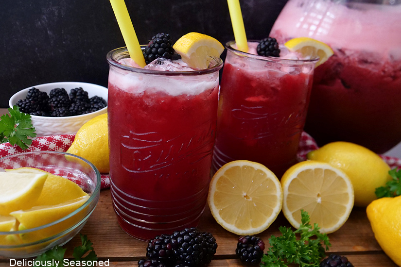 Two glasses full of blackberry lemonade with a pitcher in the background.