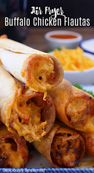 A stack of 6 chicken flautas on a white plate with blue trim with two small bowls of cheese and buffalo sauce in the background.