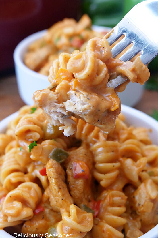 A close-up photo of a bite of chicken and pasta on a fork over a white bowl filled with pasta.