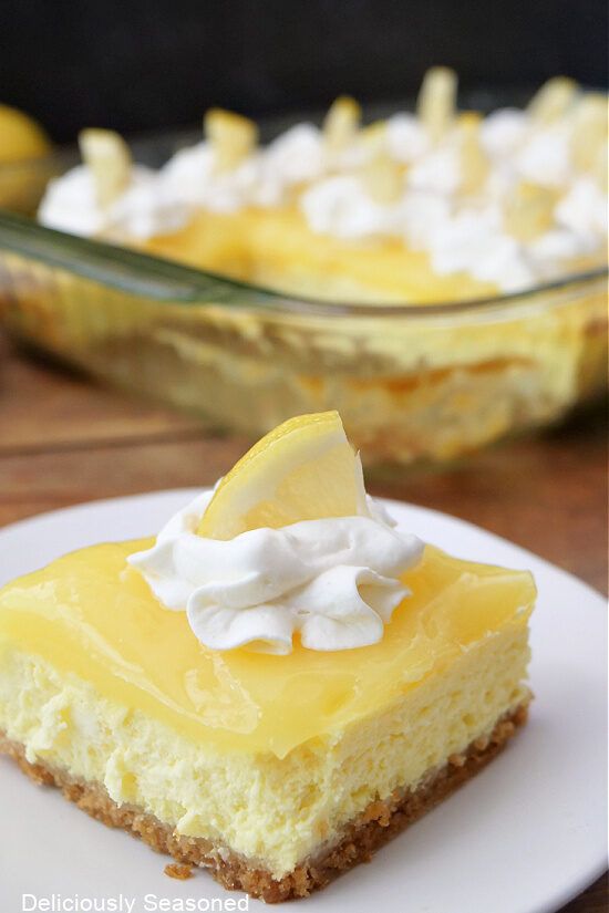 A lemon cheesecake bar on a white plate with the glass baking dish in the background.