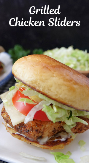 A chicken slider on a white plate showing the chicken, cheese, tomato and lettuce on a toasted slider bun.
