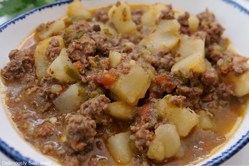 A horizontal photo of a white bowl with blue trim filled with a serving of picadillo, which is meat and potatoes.