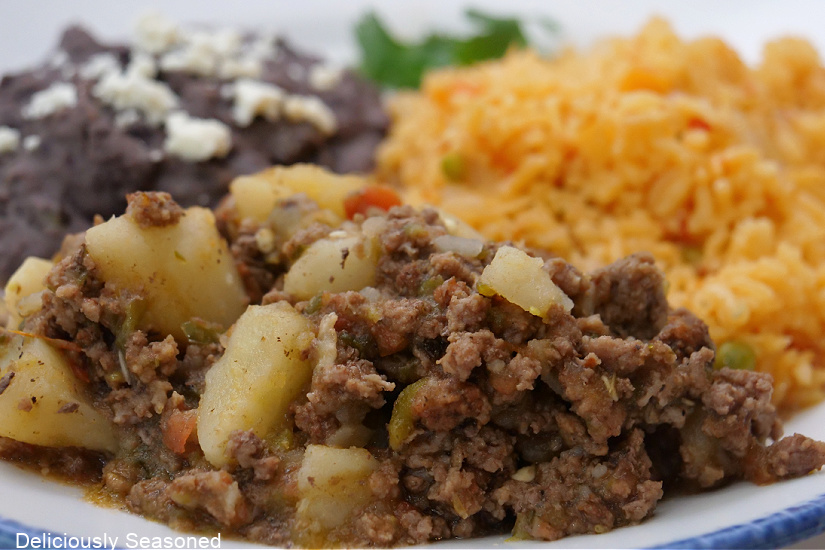 A horizontal photo of a white plate with blue trim with a serving of picadillo, Spanish rice and refried black beans.