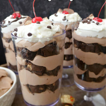 Four tall parfait glasses filled with chocolate pudding and brownie bites, topped with whipped cream and a cherry.