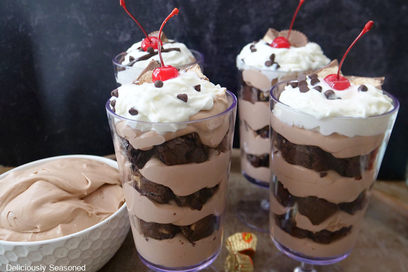 A horizontal photo of four parfait glasses and a white bowl filled with chocolate pudding and pieces of brownies.