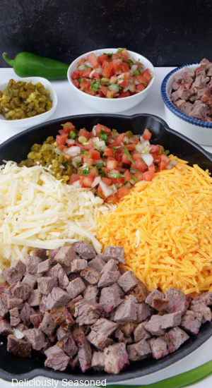 A cast iron skillet fillied with diced brisket, white and yellow shredded cheese, pico de gallo and diced jalapenos.