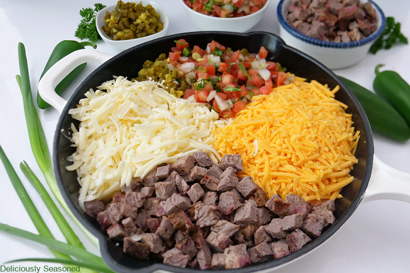 A horizontal photo of a white cast iron skillet with diced brisket, white and yellow shredded cheese and pico de gallo and diced jalapenos in it.