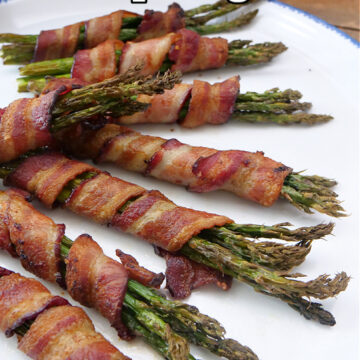 A large white plate with blue trim with 8 cooked asparagus spears wrapped in bacon.