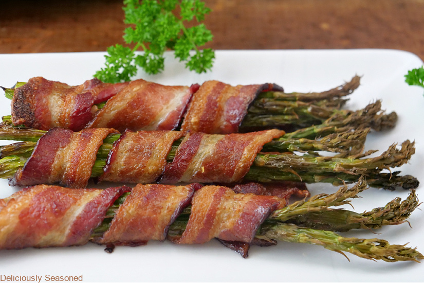 A horizontal photo of three bunches of asparagus wrapped in crispy bacon on a white plate.