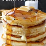 A stack of 5 pancakes on a white plate with a pad of butter on top and syrup being poured over the pancakes.