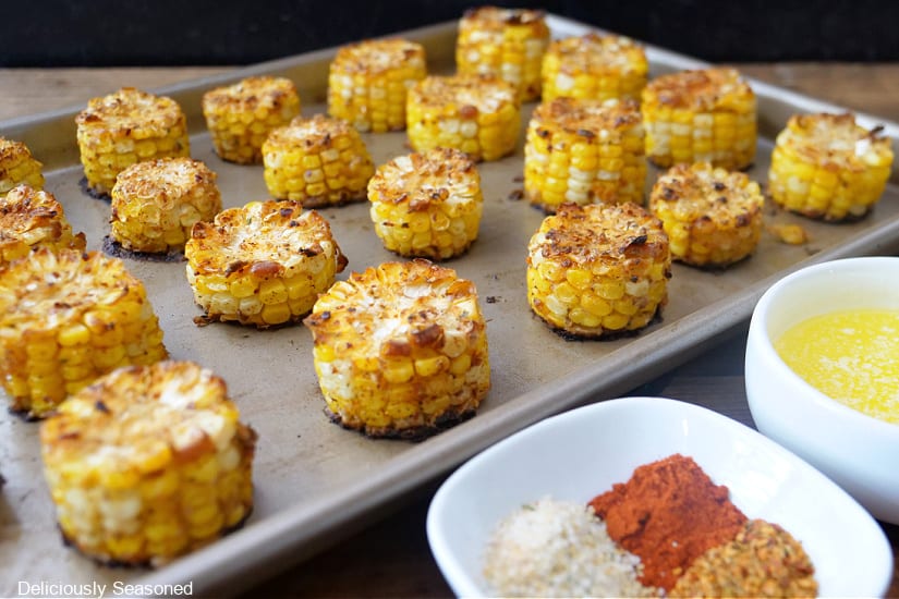 A baking sheet with mini corn on the cobs on it with melted butter and seasonings in two white bowls.