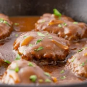 A cast iron skillet with six Salisbury steaks and gravy in it.